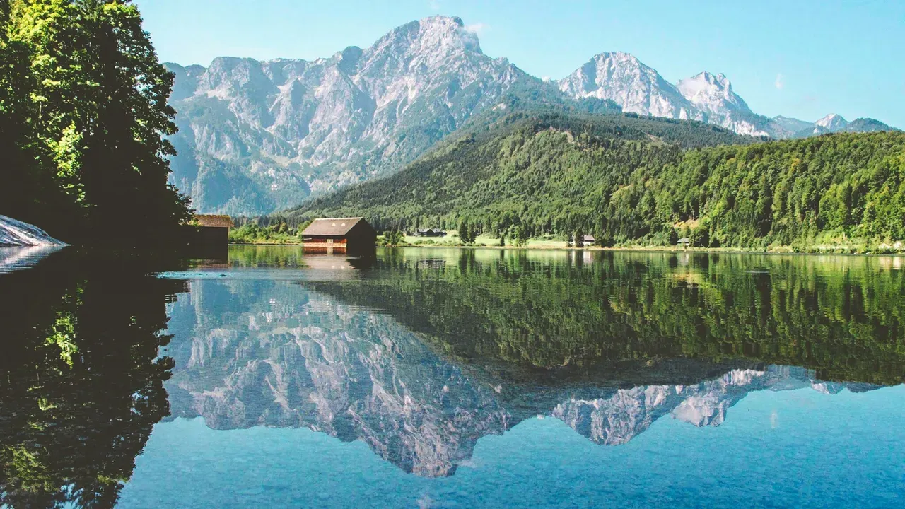 Un lac alpin paisible reflétant des versants boisés et des sommets rocheux mouchetés de neige dans les Alpes autrichiennes, avec un hangar à bateaux en bois sur la rive opposée